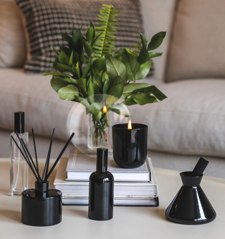 Black LUX Candle Jar with Village Craft & Candle's Aromatics collection, including the Black Boston Round Spray Bottle with Black Lid, Glass Charisma Spray Bottle with Black Lid, and Black Glossy Allure and Lavish Diffusers with Black Reeds. Product sitting on a coffee table with stacked books || Pot de bougie de luxe noir de la collection Aromatiques de Village Craft & Candle, comprenant le flacon pulvérisateur rond Boston noir avec couvercle, le flacon pulvérisateur Charisma en verre avec couvercle noir