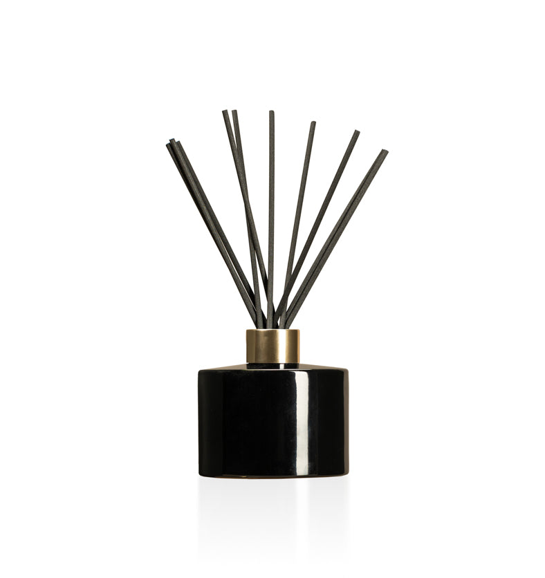Image of a stout black glass diffuser bottle with a gold collar & fiber reeds on a white background to represent Village Craft & Candle's Lavish Diffuser Bottle. || Image d'une bouteille de diffuseur en verre noire trapue avec un col doré et des tiges en fibre sur un fond blanc pour représenter la bouteille de diffuseur Lavish de Village Craft & Candle.