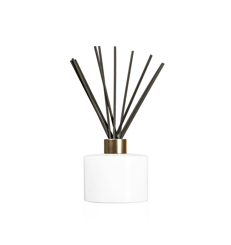 Image of a stout white glass diffuser bottle with a gold collar & fiber reeds on a white background to represent Village Craft & Candle's Lavish Diffuser Bottle. || Image d'une bouteille de diffuseur en verre blanc trapue avec un col doré et des tiges en fibre sur un fond blanc pour représenter la bouteille de diffuseur Lavish de Village Craft & Candle.