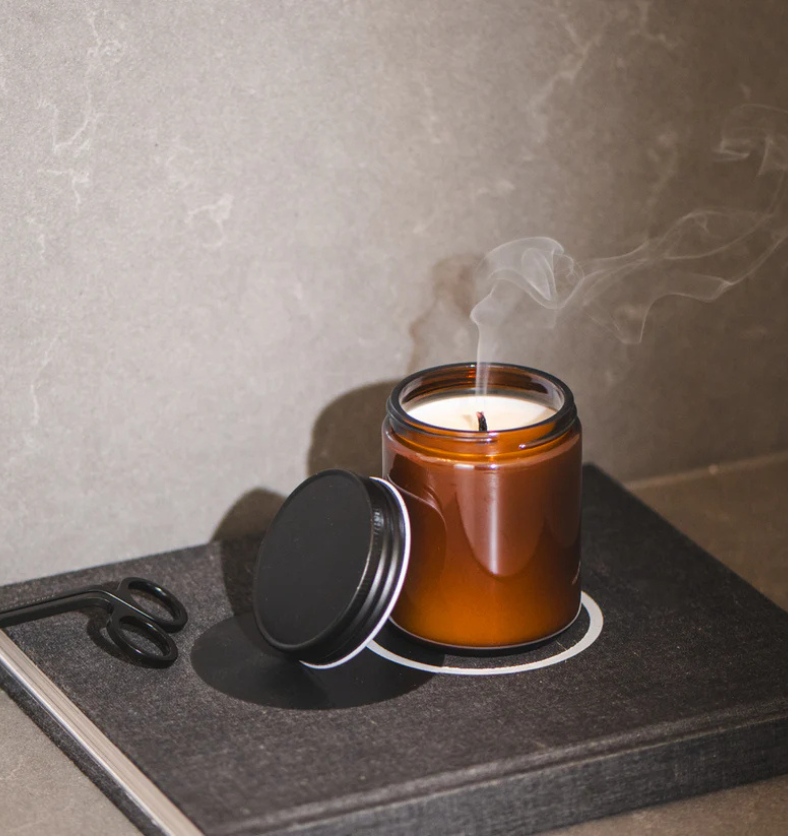 Image of an amber glass jar with matte black lid on book with wick trimmers off to side and smoke from recently blown out candle to represent Village Craft & Candle's Amber Element Straight Side Jar & Matte Black Element Lid for candle making ||
Image d’un pot en verre ambré avec un couvercle noir mat posé sur un livre, accompagné d’une pince à mèches sur le côté et de la fumée d’une bougie récemment éteinte, représentant le pot Amber Element Straight Side et le couvercle Matte Black Element de Village Cr