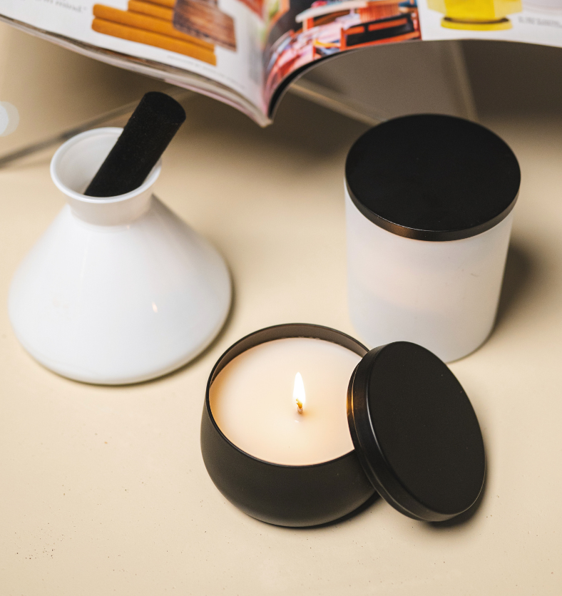 Image of a black tin candle jar and lid amongst a white aromatic diffuser and candle with a open book in the background to represent Village Craft & Candle's Black Radiance Candle Tin for candle making || Image d’un pot à bougie en étain noir avec son couvercle, contenant une bougie allumée, entouré d’un diffuseur aromatique blanc et d’une autre bougie blanche, avec un livre ouvert en arrière-plan, représentant le pot à bougie Noir Radiance de Village Craft & Candle pour la fabrication de bougies.