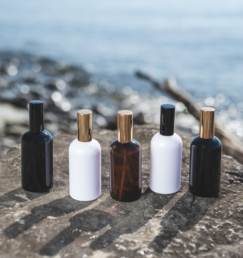 Image of black, white and amber spray bottles with gold and black lids to represent Village Craft & Candle's Boston Round Bottles from the Aromatics Collection. Bottles are on a rock with a beach in the background. || Image de vaporisateurs noir, blanc et ambré avec couvercles dorés et noirs pour représenter les bouteilles rondes Boston de la Collection Aromatics de Village Craft & Candle. Sur un rocher avec une plage en arrière-plan