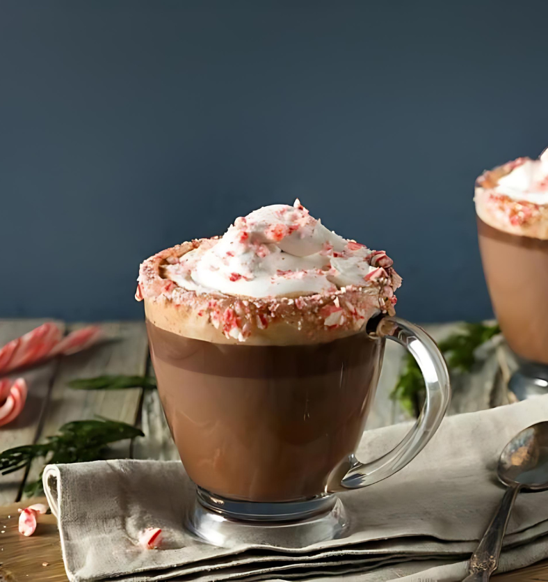Frothy mochaccino coffee in a glass mug topped with whipped cream and crushed candy cane pieces as a visual representation of Candy Cane Mochaccino Fragrance Oil available at Village Craft and Candle || Café mochaccino mousseux dans une tasse en verre garnie de crème fouettée et de morceaux de canne en bonbon écrasés comme représentation visuelle de l'huile parfumée Candy Cane Mochaccino disponible chez Village Craft and Candle