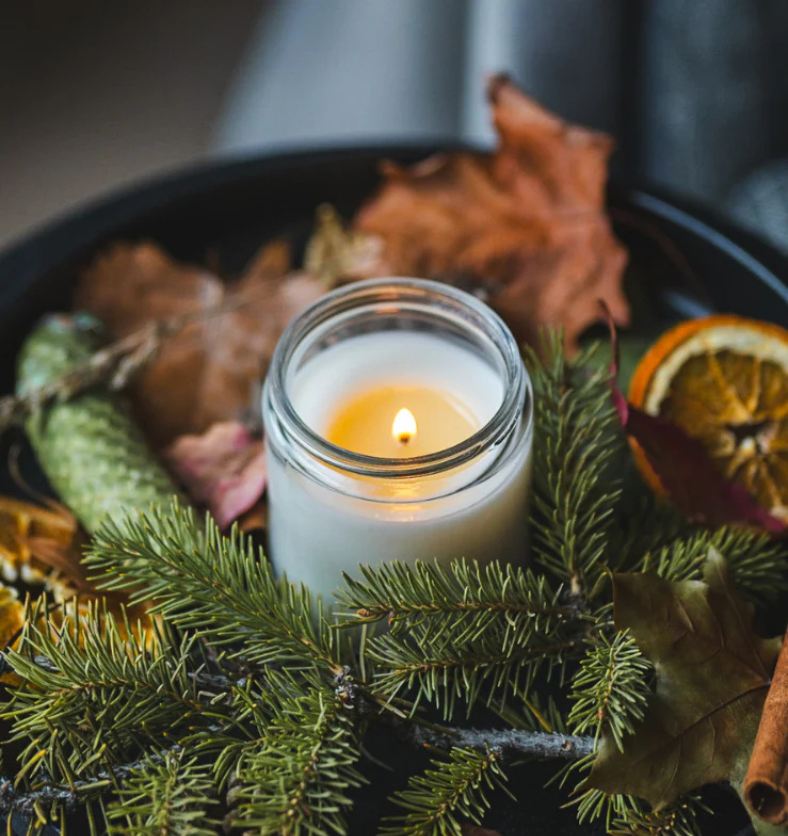 Image of a clear glass jar with lit candle amongst evergreen greenery, leaves and pinecones to represent Village Craft & Candle's Flint Clear Element Straight Side Jar for candle making || Image d’un pot en verre transparent contenant une bougie allumée, entouré de feuillage persistant, de feuilles et de pommes de pin, représentant le pot Flint Clear Element Straight Side de Village Craft & Candle pour la fabrication de bougies.