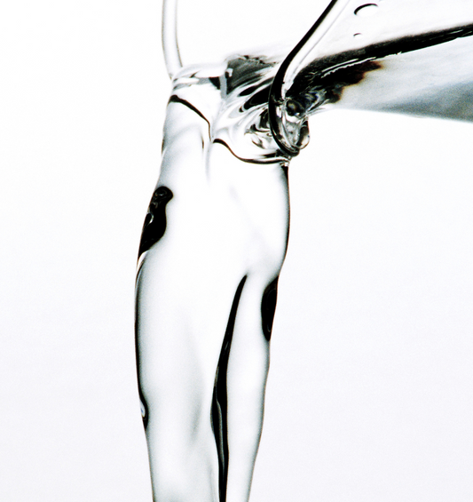 Image en gros plan d'un liquide clair versé du bec d'un récipient en verre, représentant la base de diffuseur à roseaux de Village Craft & Candle.