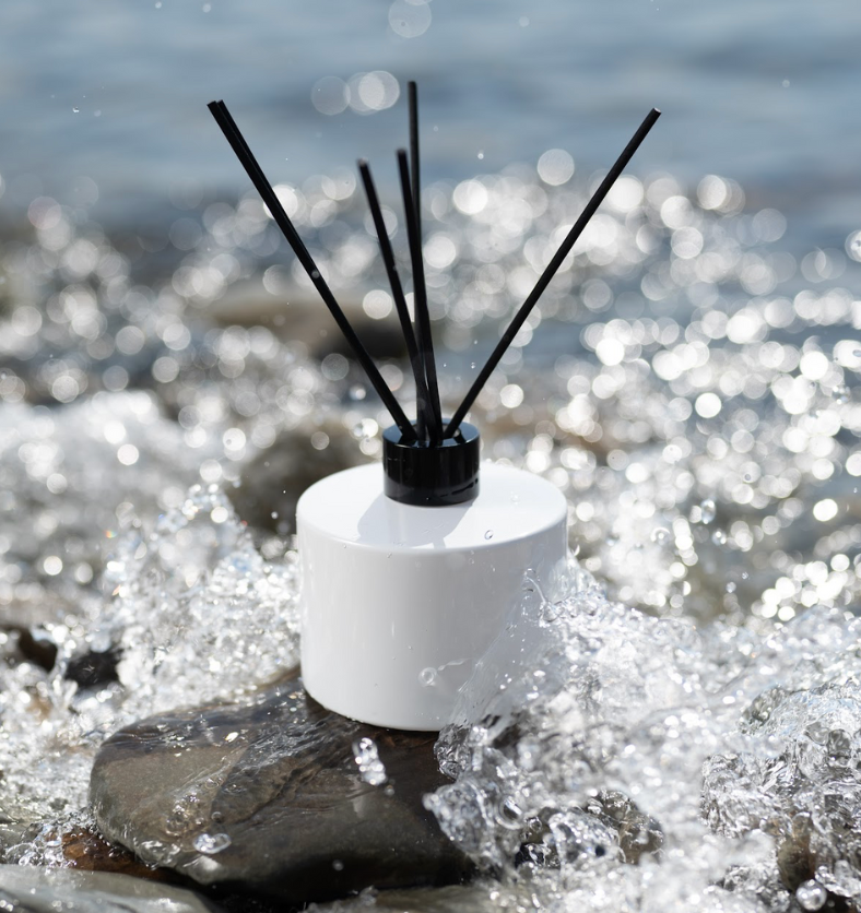 Image of Village Craft & Candle's 7oz Lavish Diffuser Bottle in White with a black collar & regular black fiber reeds sitting on a river rock being splashed by water || Image du flacon diffuseur de 7 oz Lavish de Village Craft & Candle en blanc avec un collier noir et des bâtonnets en fibres noires réguliers, posé sur un rocher de rivière éclaboussé par de l'eau.