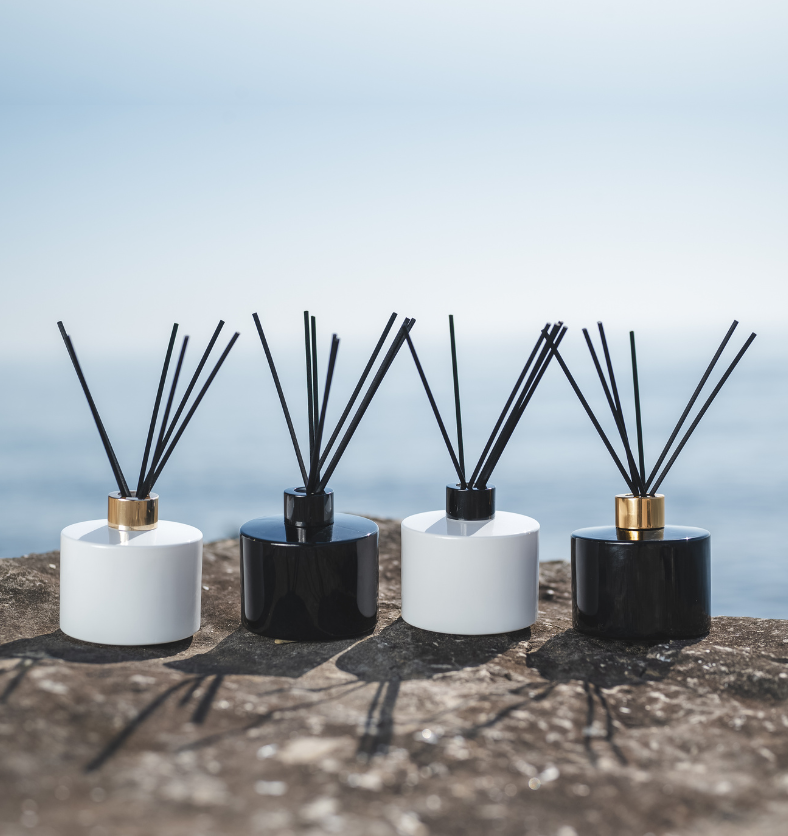 Image of black and white diffusers with gold and black lids to represent Village Craft & Candle's Lavish Diffuers from the Aromatics Collection. Diffusers are on a rock with a water in the background. || Image de diffuseurs noirs et blancs avec des couvercles dorés et noirs pour représenter les diffuseurs Lavish de Village Craft & Candle de la collection Aromatiques. Les diffuseurs sont posés sur un rocher avec de l'eau en arrière-plan.
