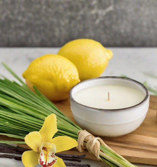 Bougie blanche à côté d'un paquet d'herbe, de citrons et d'une orchidée jaune comme représentation visuelle de l'huile parfumée à la citronnelle disponible chez Village Craft and Candle