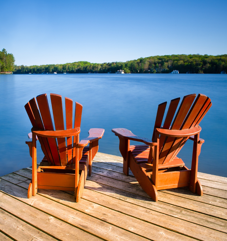 Two muskoka chairs on a fair blue lake in cottage country as a visual representation of Muskoka Lodge Fragrance Oil available at Village Craft and Candle || Deux chaises Muskoka sur un lac bleu clair dans une région de chalets comme représentation visuelle de l'huile parfumée Muskoka Lodge disponible chez Village Craft and Candle