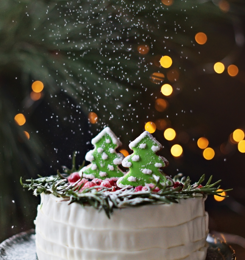 Powdered sugar falling on green gingerbread cookies atop a white cake with yellow and gold lights in the background as a visual representation of Sugared Spruce Fragrance Oil available at Village Craft and Candle || Sucre en poudre tombant sur des biscuits au pain d'épice vert au sommet d'un gâteau blanc avec des lumières jaunes et dorées en arrière-plan comme représentation visuelle de l'huile parfumée d'épicéa sucré disponible chez Village Craft and Candle
