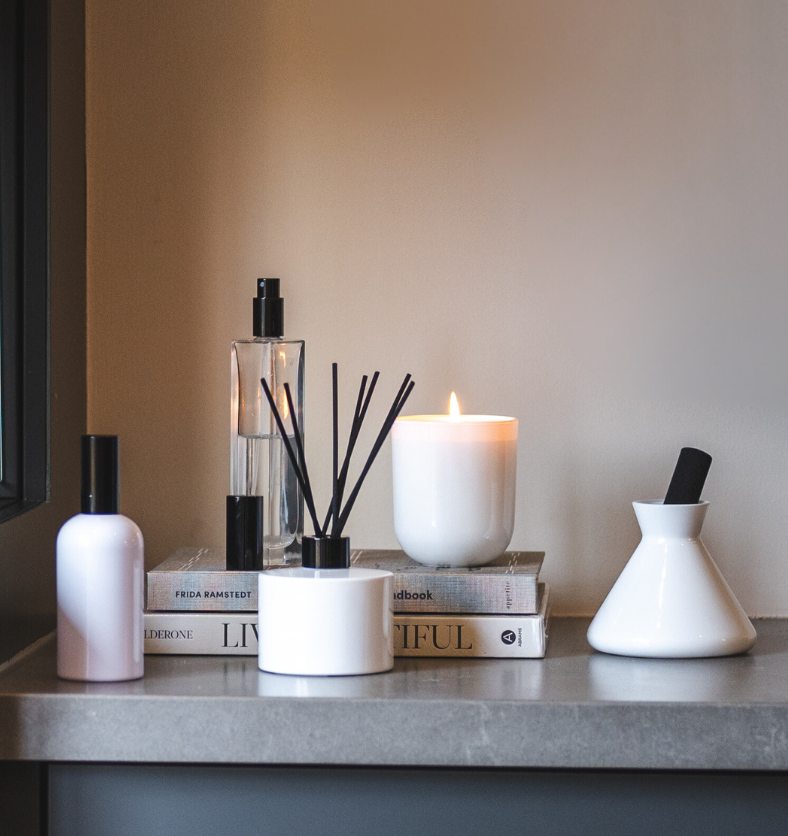 Village Craft & Candle's Charisma Glass Bottle Room Spray with Black Lid, Lavish White Glossy Diffuser, White Boston Round Room Spray with Black Lid, Allure Diffuser with black reed and White Glossy Terra Jar. Lit candle and products on stacked books on a counter. || Pulvérisateur d'ambiance Charisma avec couvercle noir, diffuseur Lavish blanc brillant, pulvérisateur rond Boston blanc avec couvercle noir, diffuseur Allure avec tiges noires et pot Terra blanc brillant