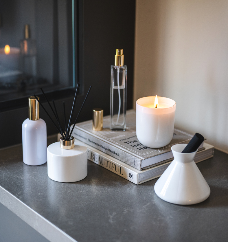 Village Craft & Candle's Aromatics Collection, featuring a lit White Glossy Terra Jar candle, Charisma Glass and White Boston Round Room Sprays with gold lids Lavish Diffuser and Allure Diffuser with black reeds on a counter atop stacked books by a fireplace || Collection Aromatics de Village Craft & Candle comprenant une bougie allumée dans un pot Terra blanc brillant des vaporisateurs de chambre Charisma en verre et White Boston Round avec des couvercles dorés un diffuseur Lavish et un diffuseur Allure