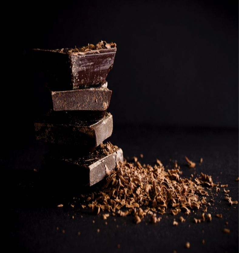Four pieces of dark chocolate stacked on top of each other against a black background with a pile of chocolate shavings spread before it as a visual representation of Dark Chocolate Fragrance Oil available at Village Craft and Candle || Quatre morceaux de chocolat noir empilés les uns sur les autres sur un fond noir avec une pile de copeaux de chocolat étalés devant eux comme représentation visuelle de l'huile parfumée au chocolat noir disponible chez Village Craft and Candle.