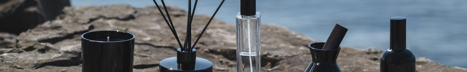 black diffuser and room spray collection with black candle lined up horizontally on edge of rock by a lake