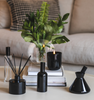 Black LUX Candle Jar with Village Craft & Candle's Aromatics collection, including the Black Boston Round Spray Bottle with Black Lid, Glass Charisma Spray Bottle with Black Lid, and Black Glossy Allure and Lavish Diffusers with Black Reeds. Product sitting on a coffee table with stacked books ||  Pot de bougie de luxe noir de la collection Aromatiques de Village Craft & Candle, comprenant le flacon pulvérisateur rond Boston noir avec couvercle, le flacon pulvérisateur Charisma en verre avec couvercle noir