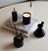 Black LUX Candle Jar with Village Craft & Candle's Aromatics collection, including the Boston Round Spray Bottle, Glass Charisma Spray Bottle with Gold Lid, and Black Glossy Allure and Lavish Diffusers with Black Reeds. Product sitting on a coffee table with stacked books || Pot de bougie de luxe noir avec la collection Aromatiques de Village Craft & Candle, comprenant le flacon Boston, le flacon Charisma en verre avec couvercle doré, et les diffuseurs Allure et Lavish noirs avec tiges