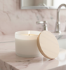 A lit candle in a 15oz Matte White LUX Jar with a White Pine lid resting next to it, placed on a pink marble bathroom countertop. || Une chandelle allumée dans un Pot LUX 15oz Blanc Mat avec un couvercle en Pin Blanc posé à côté, sur un comptoir de salle de bain en marbre rose.