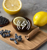 Lemon, cinnamon, and blueberries on a wood cutting board with an embossed lightning bolt as a visual representation of Agar Wood EmotiScents (Energy) Fragrance Oil available at Village Craft and Candle || Citron, cannelle et myrtilles sur une planche à découper en bois avec un éclair en relief comme représentation visuelle de l'huile parfumée Agar Wood EmotiScents (Energy) disponible chez Village Craft and Candle