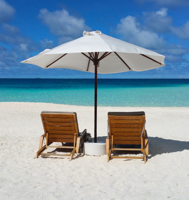 Two beach chairs on white sand beach under an umbrella as a visual representation of All Summer Long Fragrance Oil available at Village Craft and Candle || Deux chaises de plage sur une plage de sable blanc sous un parasol comme représentation visuelle de l'huile parfumée All Summer Long disponible chez Village Craft and Candle