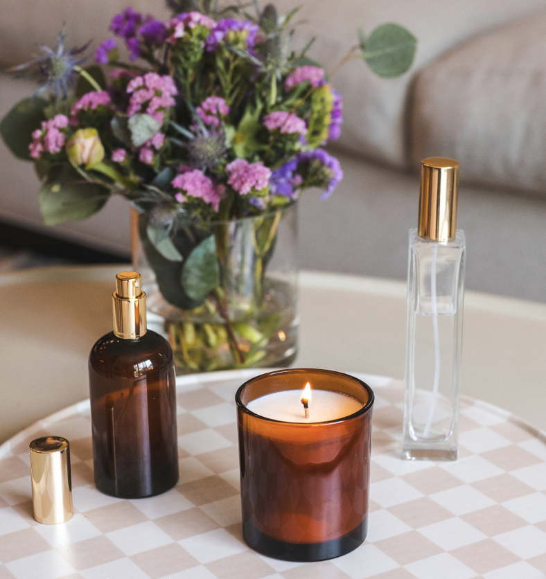 Amber Lux with Aromatic Collection room sprays. Amber Boston Round spray bottle and Charisma Glass spray bottle on a coffee table with a flower vase behind and candle lit. || Amber Lux avec la collection de vaporisateurs d'ambiance Aromatic. Vaporisateur rond Boston en ambre et vaporisateur en verre Charisma sur une table basse avec un vase à fleurs derrière et une bougie allumée