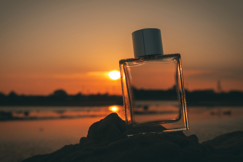 Apollo Fragrance Oil: A clear bottle of captivating fragrance with a serene beach sunset backdrop. | Huile de parfum Apollo : Un flacon transparent d'une fragrance envoûtante avec un cadre paisible d'un coucher de soleil sur la plage.