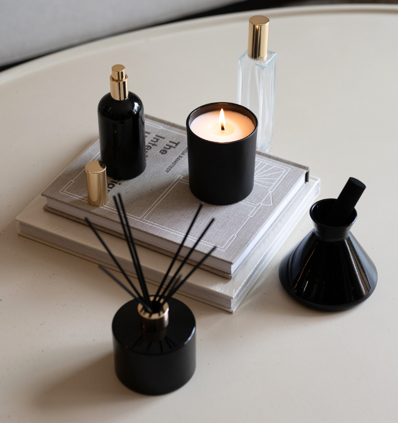 Black LUX Candle Jar with Village Craft & Candle's Aromatics collection, including the Boston Round Spray Bottle, Glass Charisma Spray Bottle with Gold Lid, and Black Glossy Allure and Lavish Diffusers with Black Reeds. Product sitting on a coffee table with stacked books. || Pot à bougie LUX noir, collection Aromatics de Village Craft & Candle. Vaporisateur rond Boston, vaporisateur en verre Charisma doré, diffuseurs Allure et Lavish noirs sur table basse avec livres empilés