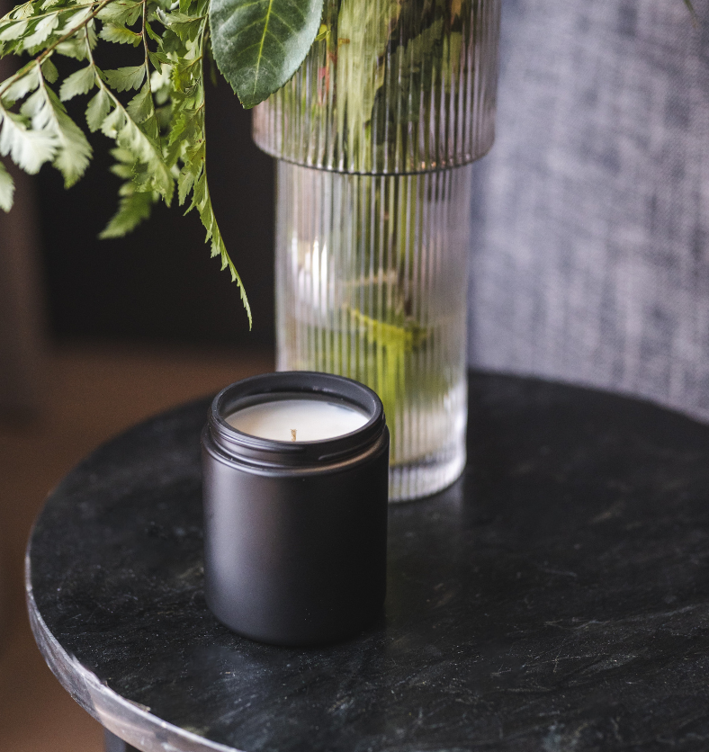 Image of a black matte jar with unlit candle on black marble table with greenery in a vase to represent Village Craft & Candle's Black Matte Element Straight Side Jar for candle making || Image d’un pot noir mat avec une bougie non allumée sur une table en marbre noir, accompagné de verdure dans un vase, représentant le pot droit Élément Noir Mat de Village Craft & Candle pour la fabrication de bougies.