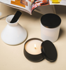 Image of a black tin candle jar and lid  amongst a white aromatic diffuser and candle with a open book in the background to represent Village Craft & Candle's Black Radiance Candle Tin for candle making || Image d’un pot à bougie en étain noir avec son couvercle, contenant une bougie allumée, entouré d’un diffuseur aromatique blanc et d’une autre bougie blanche, avec un livre ouvert en arrière-plan, représentant le pot à bougie Noir Radiance de Village Craft & Candle pour la fabrication de bougies.