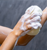 Hand using a white foamy sponge to wash an arm. || Main utilisant une éponge blanche mousseuse pour laver un bras.