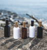 Image of black, white and amber spray bottles with gold and black lids to represent Village Craft & Candle's Boston Round Bottles from the Aromatics Collection. Bottles are on a rock with a beach in the background. || Image de vaporisateurs noir, blanc et ambré avec couvercles dorés et noirs pour représenter les bouteilles rondes Boston de la Collection Aromatics de Village Craft & Candle. Sur un rocher avec une plage en arrière-plan