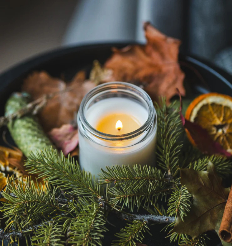 Image of a clear glass jar with lit candle amongst evergreen greenery, leaves and pinecones to represent Village Craft & Candle's Flint Clear Element Straight Side Jar for candle making ||  Image d’un pot en verre transparent contenant une bougie allumée, entouré de feuillage persistant, de feuilles et de pommes de pin, représentant le pot Flint Clear Element Straight Side de Village Craft & Candle pour la fabrication de bougies.