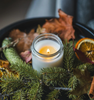 Image of a clear glass jar with lit candle amongst evergreen greenery, leaves and pinecones to represent Village Craft & Candle's Flint Clear Element Straight Side Jar for candle making ||  Image d’un pot en verre transparent contenant une bougie allumée, entouré de feuillage persistant, de feuilles et de pommes de pin, représentant le pot Flint Clear Element Straight Side de Village Craft & Candle pour la fabrication de bougies.