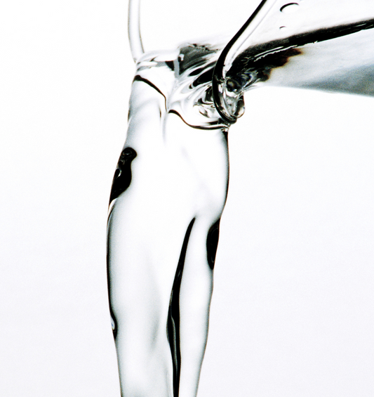 Close up image of a clear liquid pouring from the spout of a glass vessel to represent Village Craft & Candle's Reed Diffuser Base. 