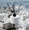 Image of Village Craft & Candle's 7oz Lavish Diffuser Bottle in White with a black collar & regular black fiber reeds sitting on a river rock being splashed by water || Image du flacon diffuseur de 7 oz Lavish de Village Craft & Candle en blanc avec un collier noir et des bâtonnets en fibres noires réguliers, posé sur un rocher de rivière éclaboussé par de l'eau. 