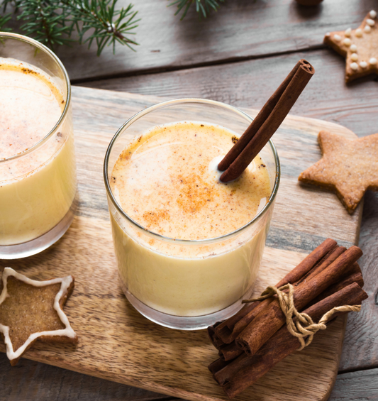 Glass of eggnog on a wood board with cinnamon and gingerbread cookies as a visual representation of Eggnog Fragrance Oil available at Village Craft and Candle 