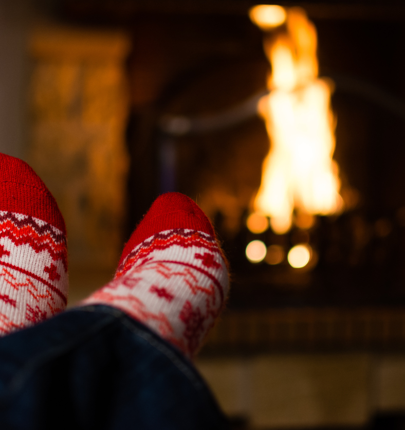 Christmas socks on feet next to a fireplace as a visual representation of Fireside Fragrance Oil available at Village Craft and Candle || Chaussettes de Noël sur pieds à côté d'une cheminée comme représentation visuelle de l'huile parfumée au coin du feu disponible chez Village Craft and Candle