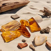 Image of pieces of amber and drift wood laying on sand to represent Village Craft & Candle's Golden Santal Fragrance Oil || Image de morceaux d'ambre et de bois flotté posés sur le sable pour représenter l'huile parfumée Golden Santal de Village Craft & Candle