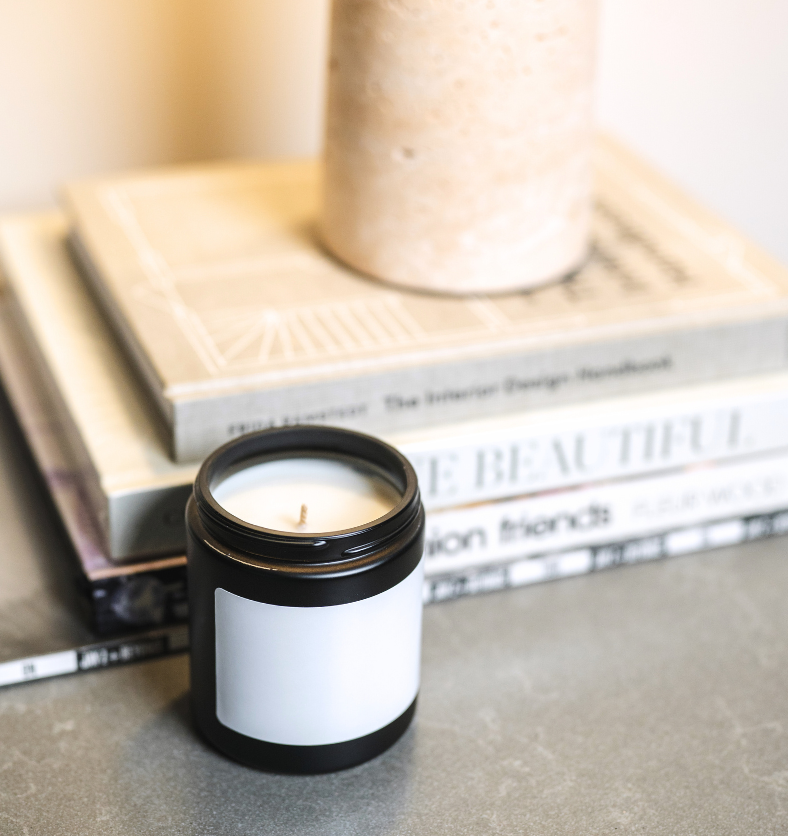 Image of an unlit candle in black matte jar with white blank label on the front and stacked books in the background to represent Village Craft & Candle's Blank 4" x 2" Credit Card Label || Image d’une bougie non allumée dans un pot noir mat avec une étiquette blanche vierge à l’avant, et des livres empilés en arrière-plan, représentant l’étiquette vierge 4" x 2" format carte de crédit de Village Craft & Candle.