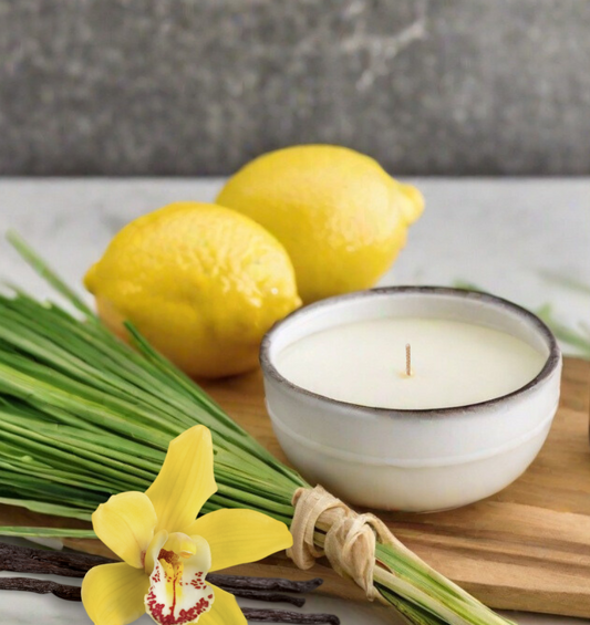  Bougie blanche à côté d'un paquet d'herbe, de citrons et d'une orchidée jaune comme représentation visuelle de l'huile parfumée à la citronnelle disponible chez Village Craft and Candle