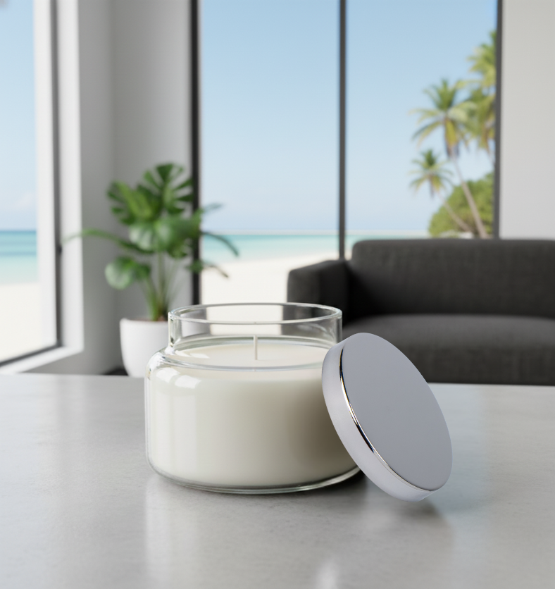 A finished 10oz Traditional Jar candle with a wick, sitting on a modern table next to its silver lid, with an ocean beach and palm trees visible through the window. || Une chandelle finie dans le Pot Traditionnel 10oz avec mèche, posée sur une table moderne à côté de son couvercle argenté, avec une plage et des palmiers visibles par la fenêtre.