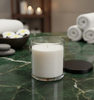 Lifestyle image of the new 8oz Madison Jar, filled as a white candle, sitting on a green marble surface in a spa setting with rolled towels and decorative stones in the background. | Image d'ambiance du nouveau pot Madison de 8 oz, rempli comme une bougie blanche, posé sur une surface en marbre vert dans un décor de spa avec des serviettes roulées et des pierres décoratives en arrière-plan.