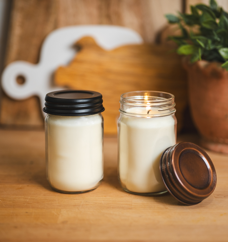 Two 12oz mason jar candles with black and copper Country lids from Village Craft & Candle, displayed on a wooden surface with a cozy background of cutting boards and a potted plant || Deux bougies en pots Mason de 12 oz avec des couvercles Country noirs et cuivrés de Village Craft & Candle, présentées sur une surface en bois avec un arrière-plan chaleureux composé de planches à découper et d'une plante en pot.