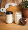 Two 12oz mason jar candles with black and copper Country lids from Village Craft & Candle, displayed on a wooden surface with a cozy background of cutting boards and a potted plant || Deux bougies en pots Mason de 12 oz avec des couvercles Country noirs et cuivrés de Village Craft & Candle, présentées sur une surface en bois avec un arrière-plan chaleureux composé de planches à découper et d'une plante en pot.