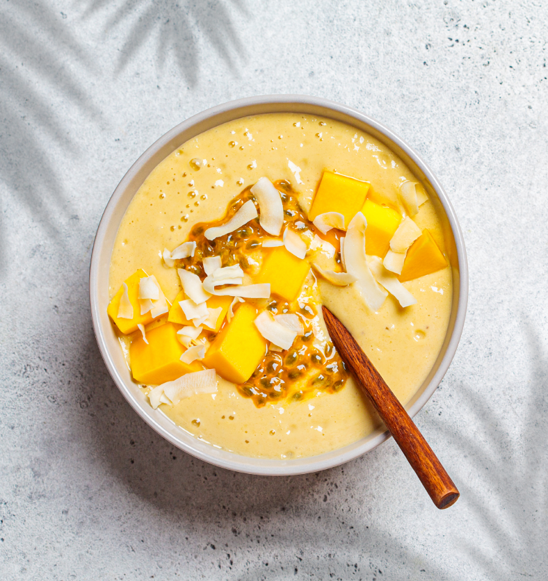 Mango smoothie bowl with wooden spoon and cut up mango pieces with coconut flakes on top as a visual representation of Mango Coconut Fragrance Oil available at Village Craft and Candle || Bol de smoothie à la mangue avec une cuillère en bois et des morceaux de mangue coupés, garnis de flocons de noix de coco, en tant que représentation visuelle de l'Huile Parfumée Mangue Noix de Coco disponible chez Village Craft and Candle.