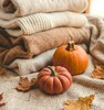 Stack of folded beige and brown sweaters with small pumpkins and leaves on a textured surface