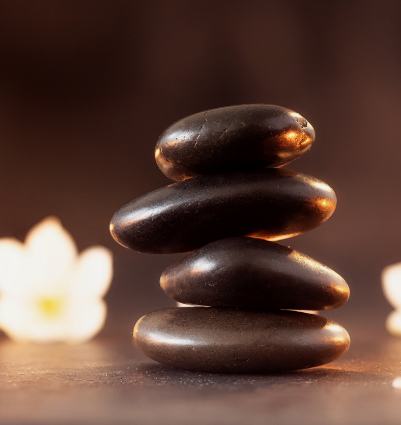 Stacked brown stones in foreground with flowers and candles as a visual representation of Spa Fragrance Oil available at Village Craft and Candle || Pierres brunes empilées au premier plan avec des fleurs et des bougies comme représentation visuelle de l'huile parfumée de spa disponible chez Village Craft and Candle