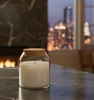 Finished candle in a 13oz apothecary jar with a cork lid sitting on a dark marble countertop. A glowing fireplace is on the left, and a blurred nighttime cityscape is visible through the window in the background. || Chandelle finie dans un pot apothicaire de 13 oz avec couvercle en liège posé sur un comptoir en marbre foncé. Une cheminée allumée est à gauche, et une ville floue illuminée la nuit est visible par la fenêtre en arrière-plan.