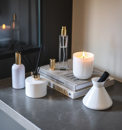 Village Craft & Candle's White Glossy Terra Jar lit candle with Aromatics Collection, including Charisma Glass Room Spray Bottle with gold lid, White Boston Round Room Spray with gold lid, Lavish Diffuser and Allure Diffuser with black reeds. Products sitting on a counter on top of stacked books by a fireplace || Bougie Terra blanche de Village Craft & Candle avec Collection Aromatics : vaporisateur Charisma en verre doré, vaporisateur rond Boston blanc doré, diffuseurs Lavish et Allure avec roseaux noirs. 