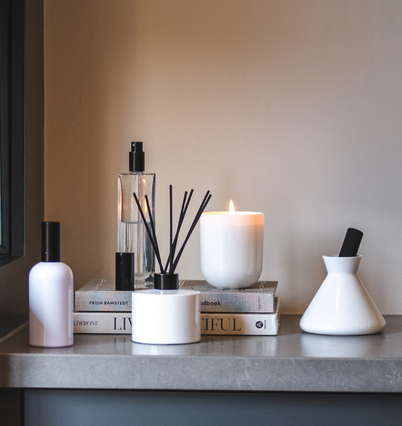 Village Craft & Candle's Charisma Glass Bottle Room Spray with Black Lid, Lavish White Glossy Diffuser, White Boston Round Room Spray with Black Lid, Allure Diffuser with black reed and White Glossy Terra Jar. Lit candle and products on stacked books on a counter. ||  Pulvérisateur d'ambiance Charisma avec couvercle noir, diffuseur Lavish blanc brillant, pulvérisateur rond Boston blanc avec couvercle noir, diffuseur Allure avec tiges noires et pot Terra blanc brillant