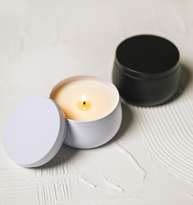 Image of a lit candle in a white tin with matching lid leaning on the side of jar and black version behind to represent Village Craft & Candle's White and Black Radiance Candle Tin for candle making || Image d’une bougie allumée dans un pot en étain blanc avec son couvercle assorti appuyé sur le côté du pot, avec la version noire en arrière-plan, représentant les pots à bougie Blanc et Noir Radiance de Village Craft & Candle pour la fabrication de bougies.